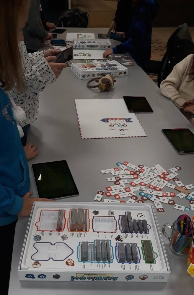 Dzieci uczestniczą w zajęciach z programowania. Stoją przy stole, na którym znajduje się gra edukacyjna Scottie Go. Za pomocą tabletów uczą się programowania.
