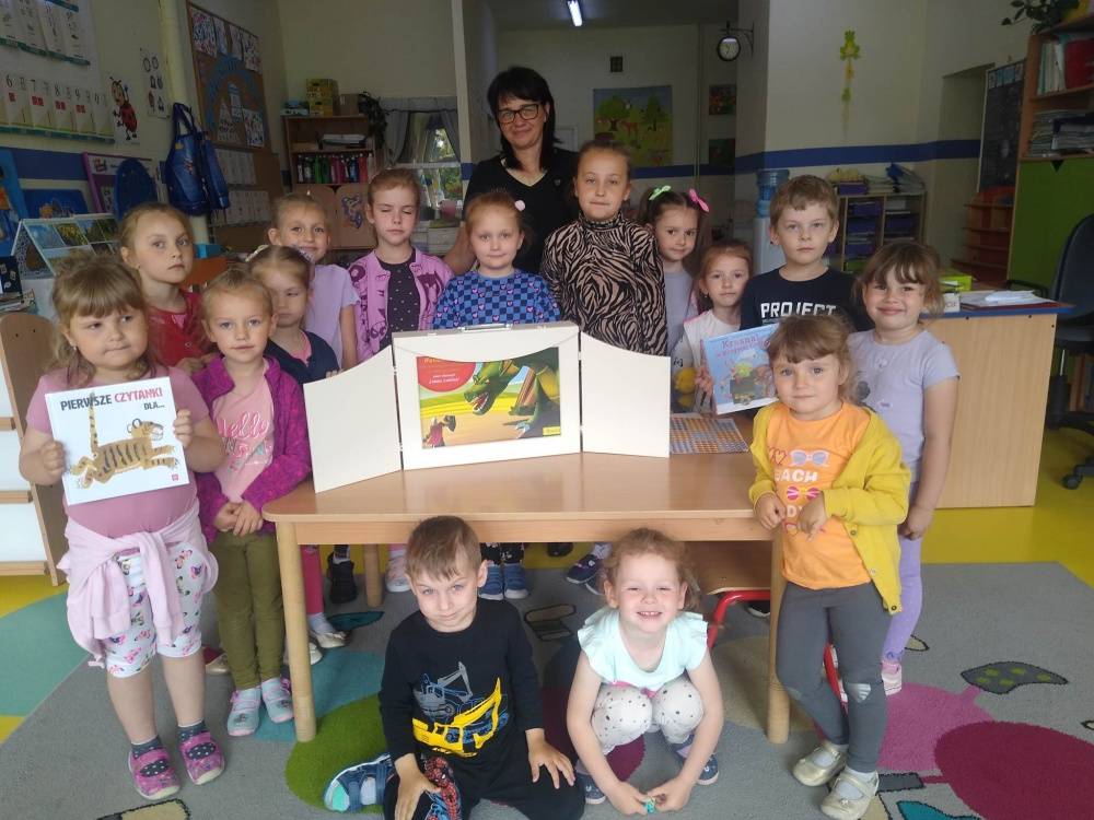 Zdjęcie grupowe dzieci w przedszkolu. Na biurku stoi biały teatrzyk Kamishibai, w której znajduje się książka pt. "Legenda o Smoku Wawelskim".