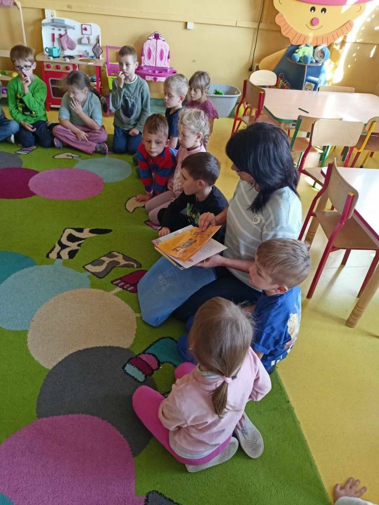 Dzieci w przedszkolu siedzą na kolorowym dywanie. Słuchają bibliotekarki, która opowiada im o bibliotece i zasadach wypożyczania książek.
