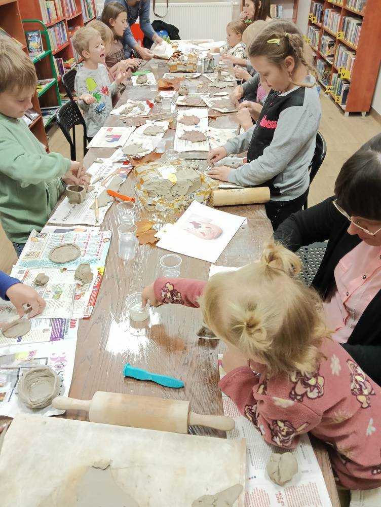 Zajęcia ceramiczne dla dzieci w bibliotece. Przy długim stole uczestnicy tworzą wyroby z gliny z jesiennymi motywami.
