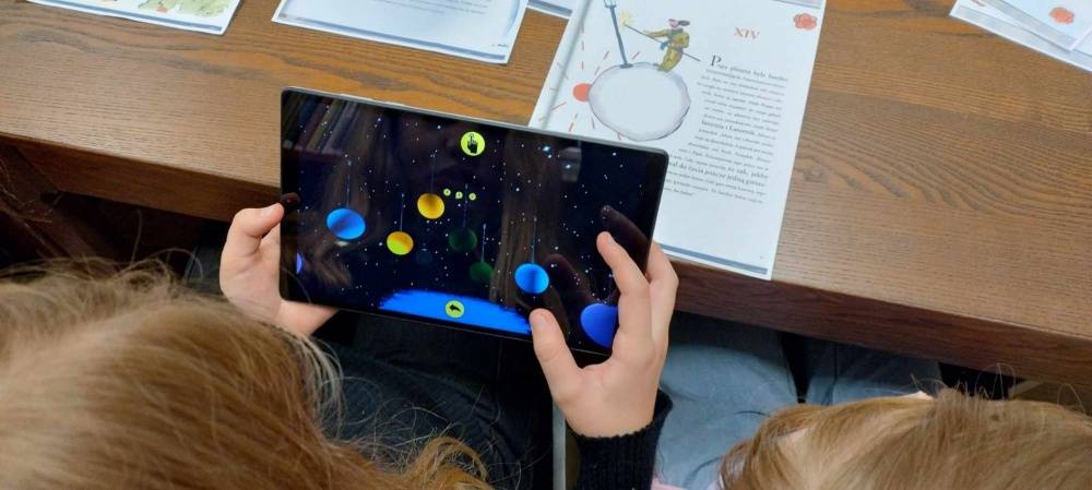 Młoda osoba trzyma w rękach tablet, za pomocą którego poznaje technologię rozszerzonej rzeczywistości. Na ekranie widoczna galaktyka i krążące planety.