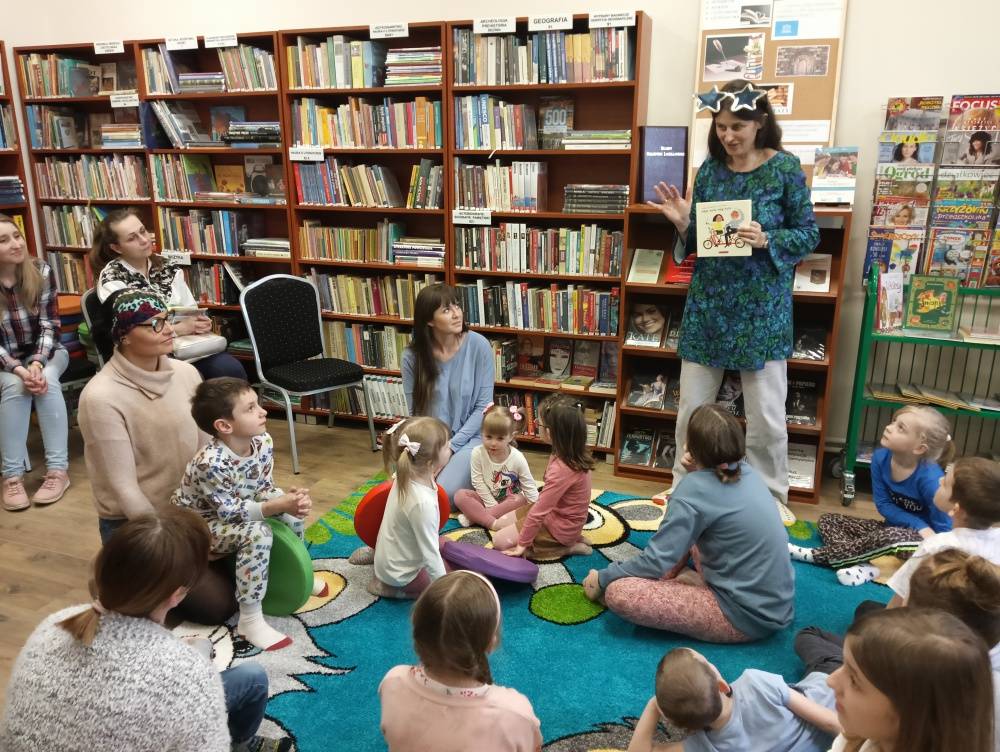 Warsztaty dla rodzin w bibliotece na temat czytania wrażeniowego. Dzieci wraz z rodzicami siedzą na niebieskim dywanie i słuchają wypowiedzi zaproszonej pisarki.