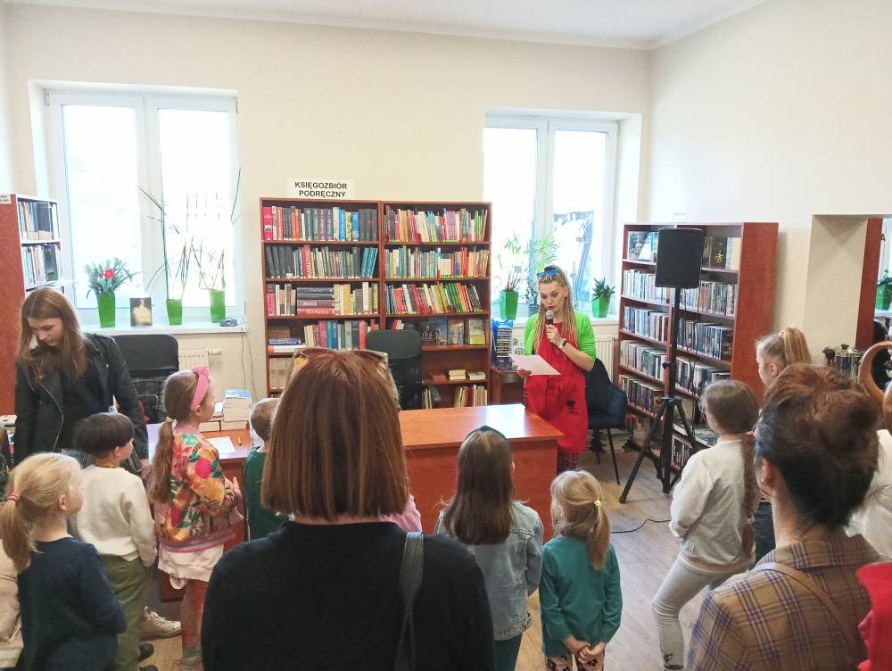 Dzieci wraz z opiekunami, zgromadzeni w bibliotece, słuchają wiersza czytanego przez kobietę, na temat traktowania i karmienia alpak.