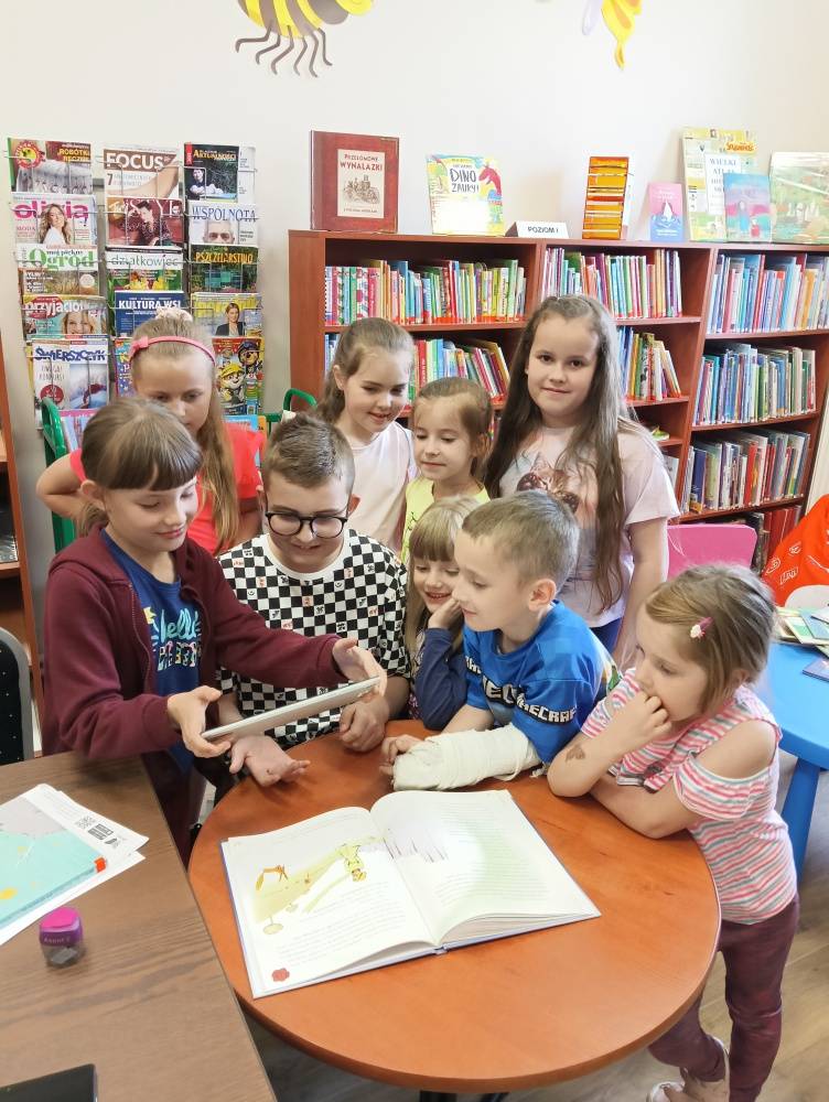 Dzieci znajdują się w bibliotece, oglądają ilustracje w książce z wykorzystaniem tableta i aplikacji AR.