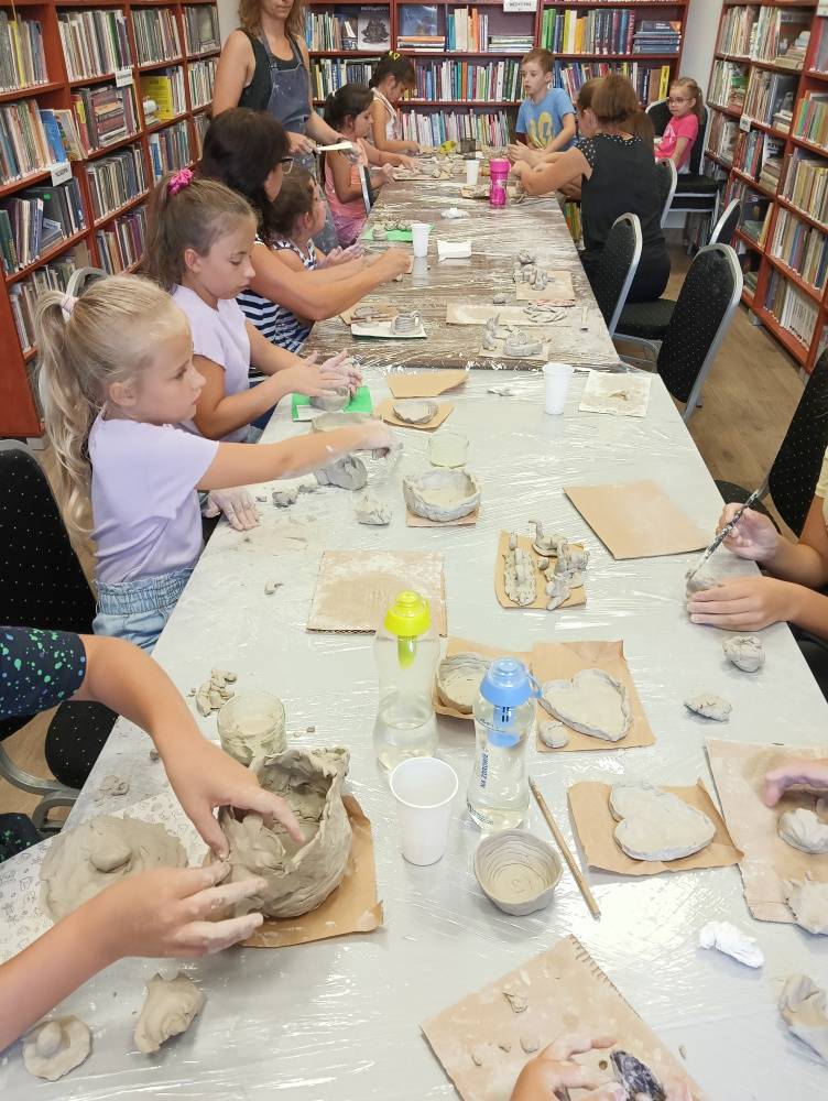 Warsztaty ceramiczne w bibliotece. Dzieci siedzą przy długim stole i wykonują różne przedmioty z gliny.