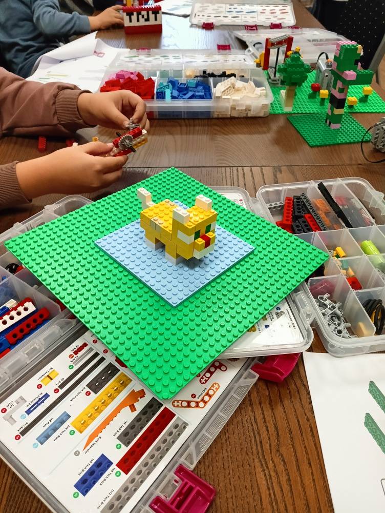 Na stole znajdują się pudełka z klockami Lego. Dzieci układają z nich różne modele. Na środku zdjęcia żółty kotek z klocków na zielonej podstawce.
