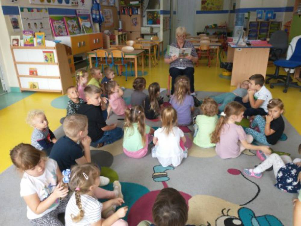 Dzieci w przedszkolu siedzą na dywanie, słuchają czytanej przez bibliotekarkę książki.