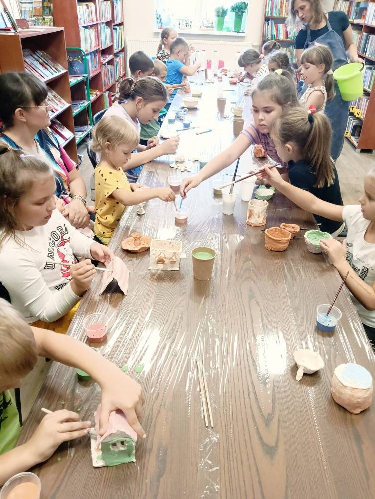 W bibliotece dzieci malują swoje wyroby z gliny. Siedzą przy długim, brązowym stole, na którym znajdują się kubeczki z farbami.