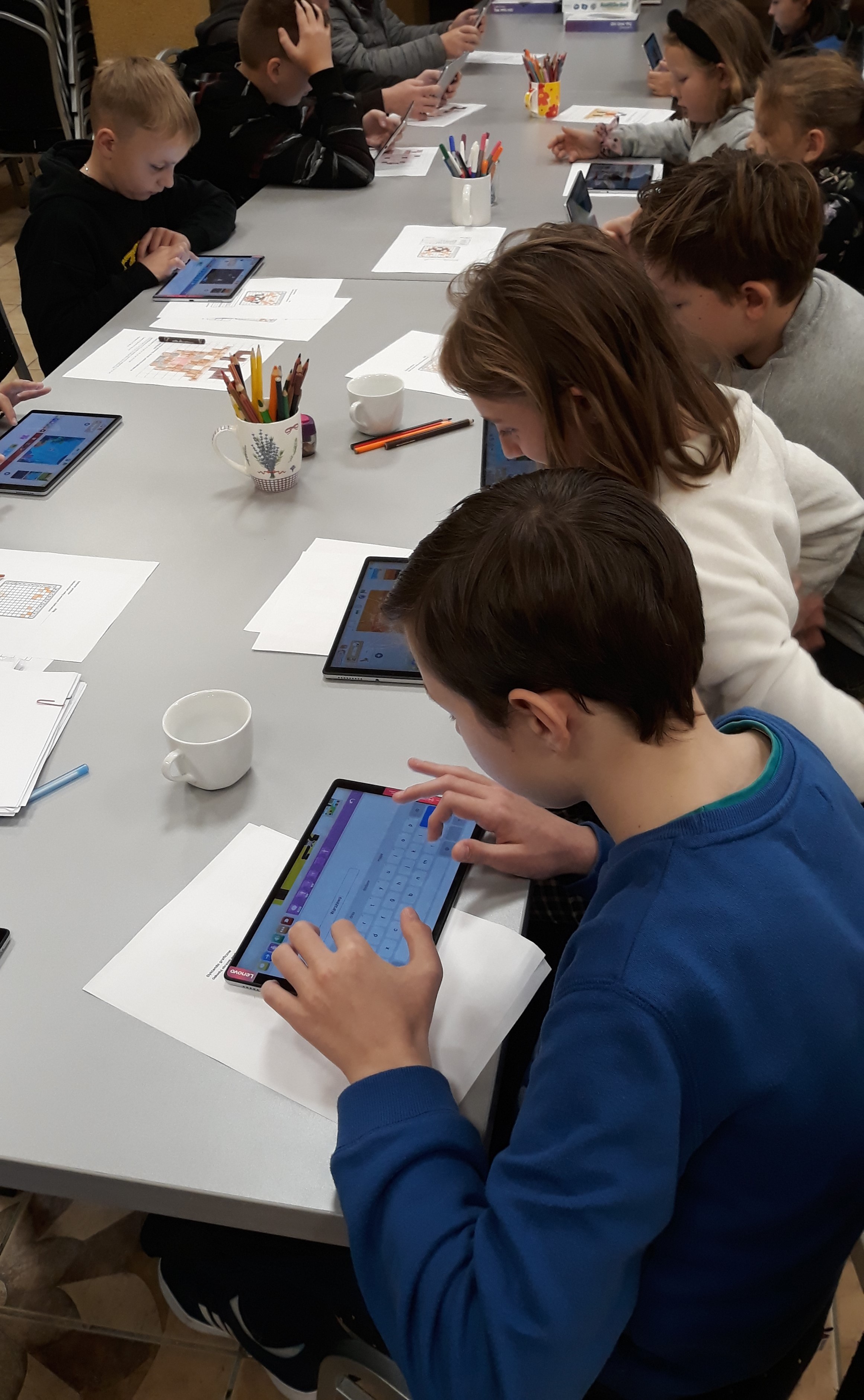 Dzieci siedzą przy stole, poznają na tablecie aplikacje do nauki programowania. Na stole widoczne kartki i przybory do rysowania.
