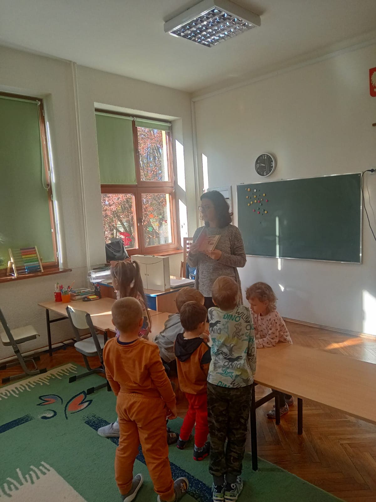 W klasie, małe dzieci stoją przy biurku i oglądają książeczkę prezentowaną przez kobietę. W tle zielona szkolna tablica.