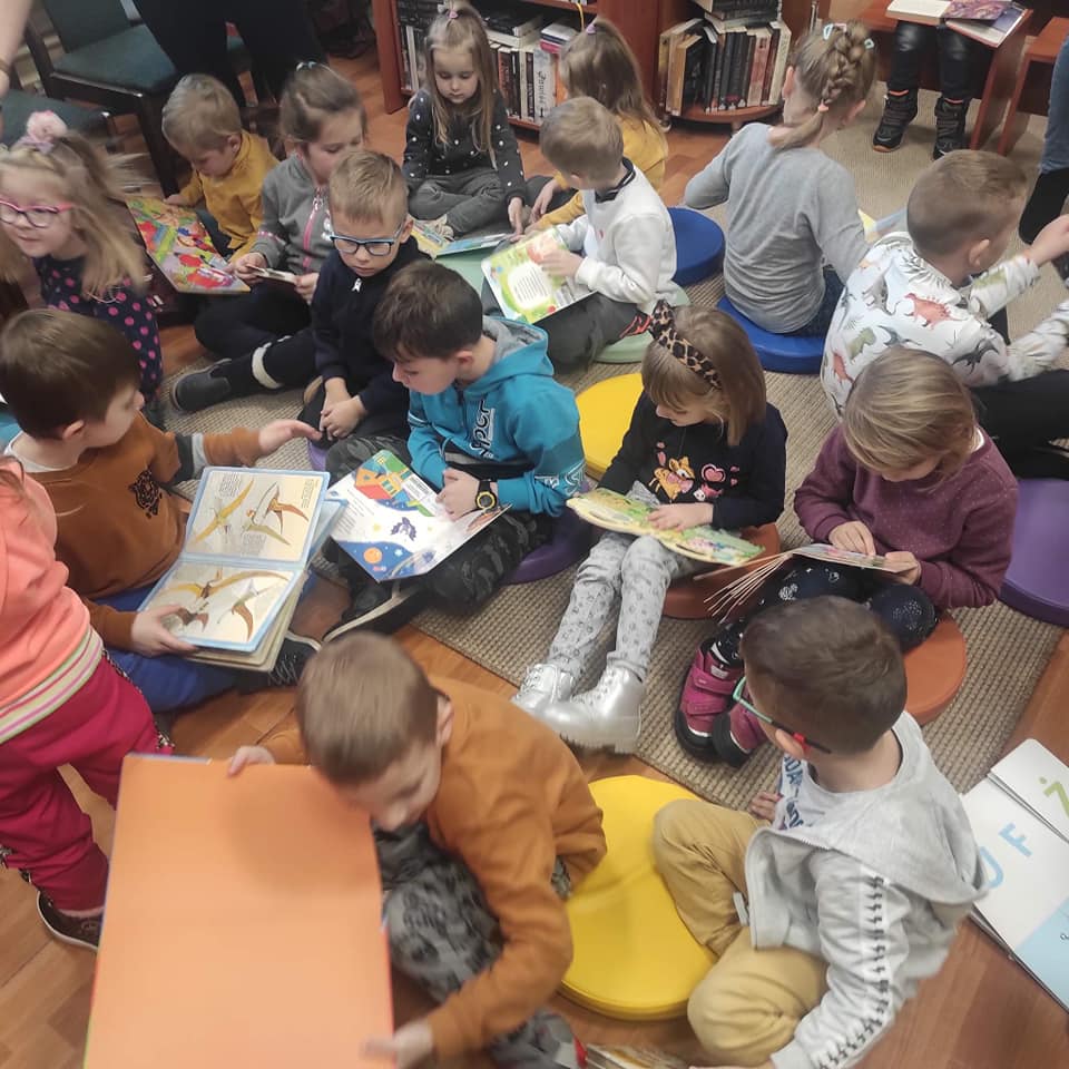 Dzieci siedząc na dywanie oglądają przygotowane przez bibliotekarzy kolorowe książeczki.